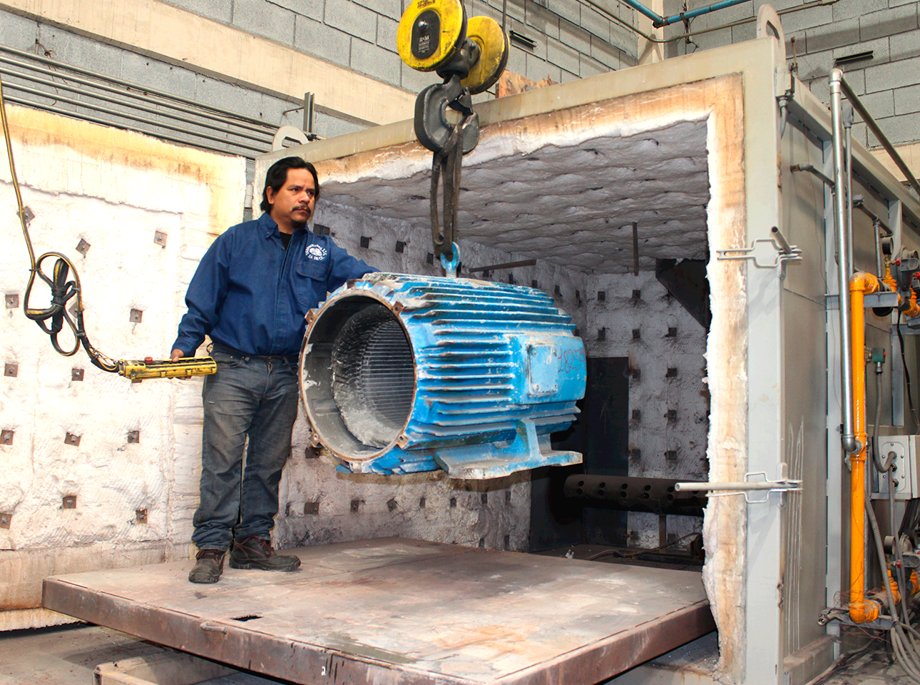 Técnico de mantenimiento industrial manipulando una carcasa de motor eléctrico con grúa en horno de tratamiento térmico.